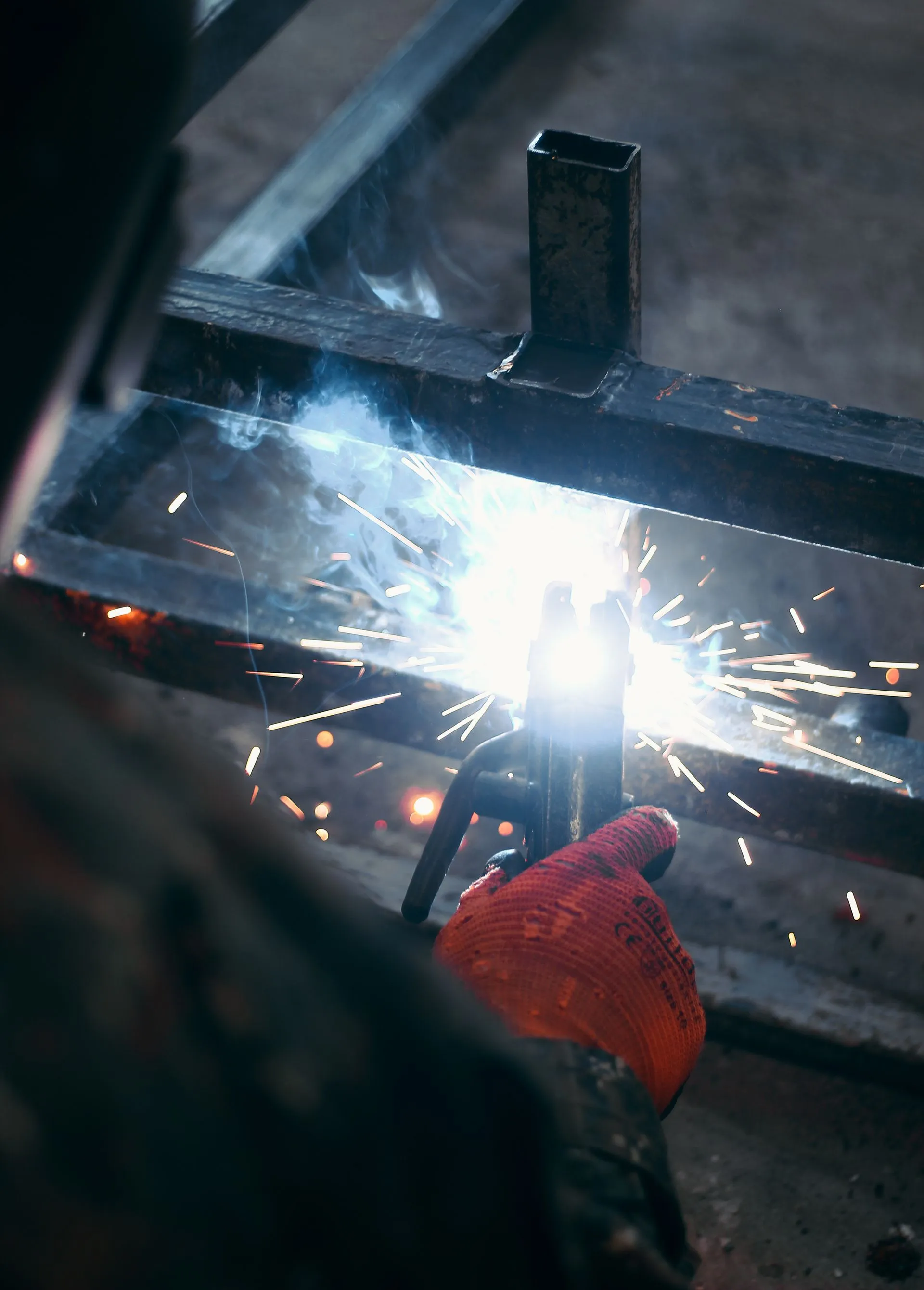 A welder gripping an electrode holder mid-weld on a steel frame, sparks flying