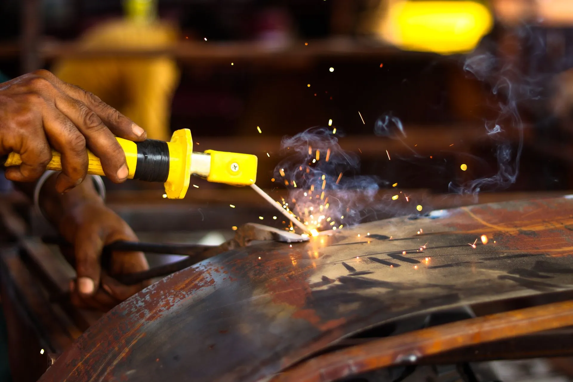 A bare hand gripping a bright yellow electrode holder with the rod clearly visible, sparks flying in a daylit workshop